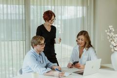 2 women sitting at a table, looking at a laptop, 1 woman standing behind them holding a coffee cup